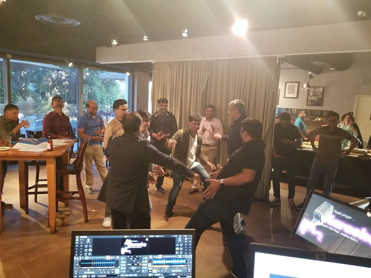 A group of people socialize and dance in a casual indoor setting, with tables, computers, and food visible in the background.