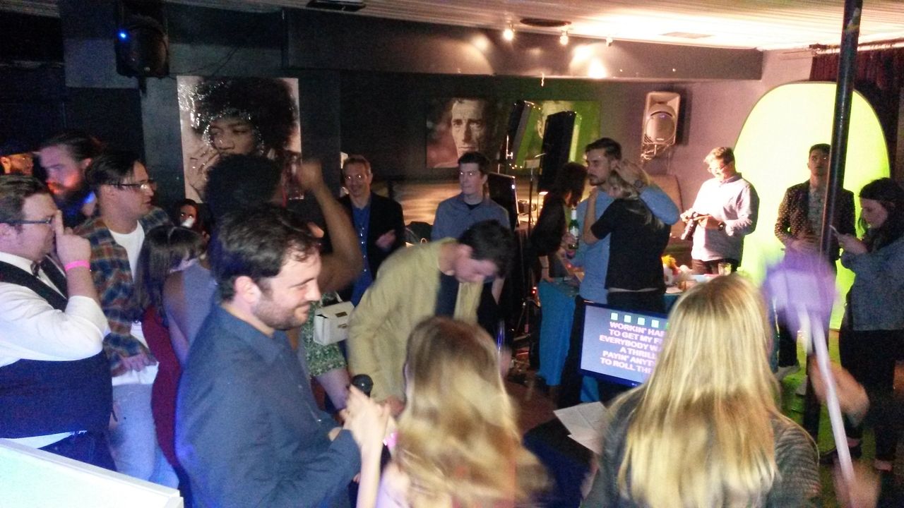 A group of people gather and socialize in a dimly lit room with a green screen and karaoke machine, while a few hold microphones and sing.