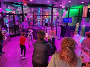 Children dance at a birthday party in a room with purple lighting, balloons, and a DJ booth while adults watch and take photos.