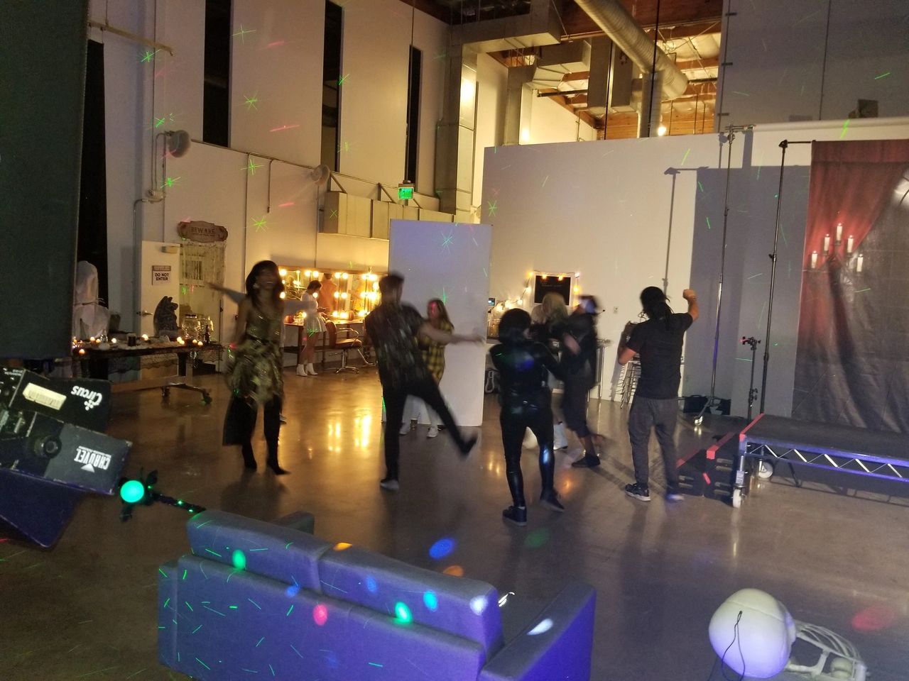 People are dancing in a dimly lit room with scattered furniture and colored lights; a stage and dressing area are visible in the background.