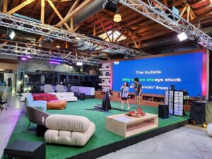 Two people stand on a stage with large karaoke lyrics displayed on a screen behind them in a spacious, modern, loft-style room with casual seating and exposed beams.