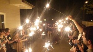 People form two lines holding sparklers at night while a couple dances in the middle of a pathway outside a building.