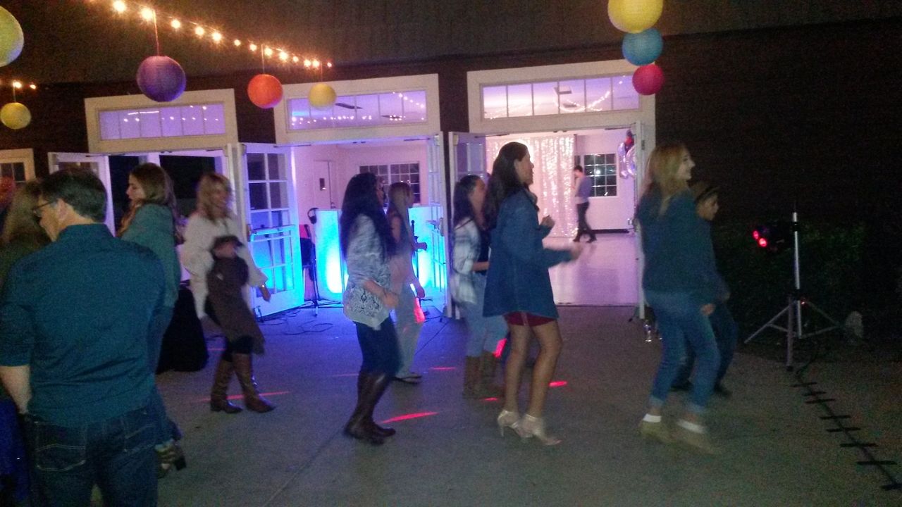 A group of people dance together on a patio at night under string lights and colorful paper lanterns, with a building and open doors in the background.