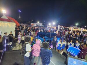 Large crowd at a nighttime outdoor event with tents, a blue car, performers on stage, and various people walking and gathering under bright lights.