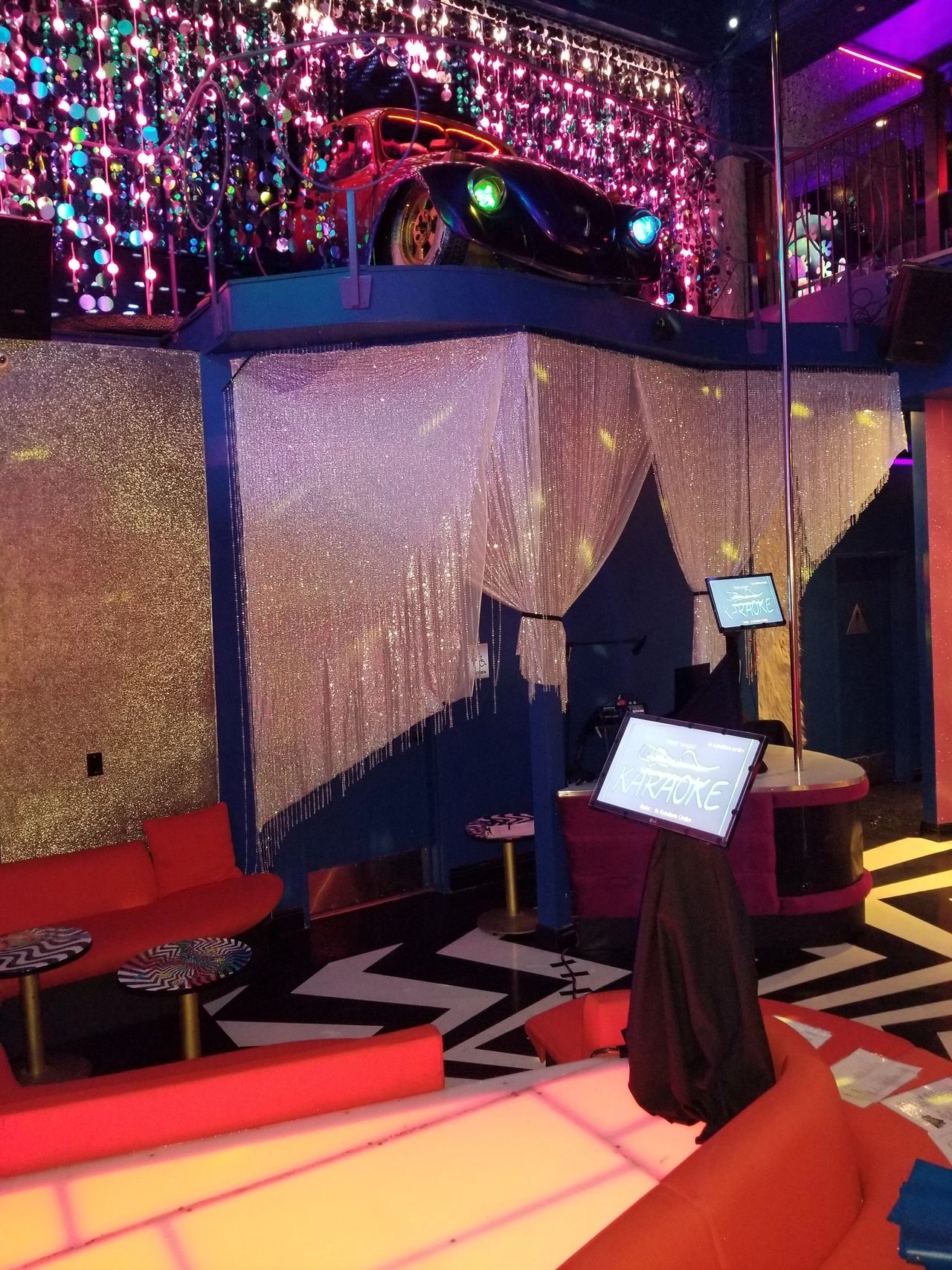 A brightly lit karaoke lounge with red seating, a stage, a karaoke machine, glittery decor, and a car displayed on a mezzanine level above.