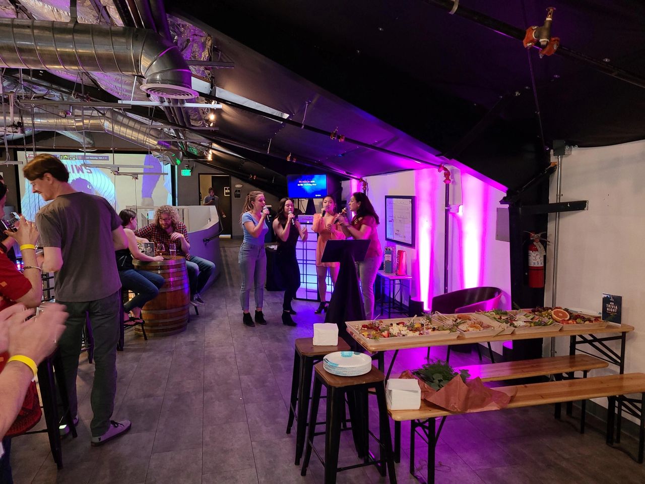 People socialize in a dimly lit venue with purple lighting; three women sing by a wall, and a table in the foreground holds food and plates.