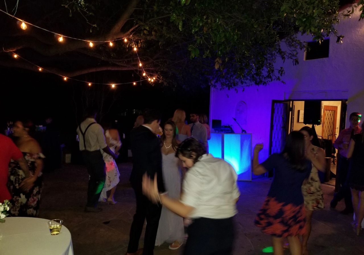 People dance outdoors at night under string lights, with a lit DJ booth and a white building in the background. Drinks and tables are visible in the scene.