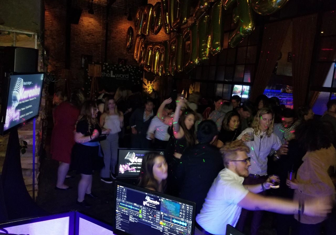 A crowd of people dance and socialize at a dimly lit indoor party with gold balloons and a karaoke setup visible in the foreground.
