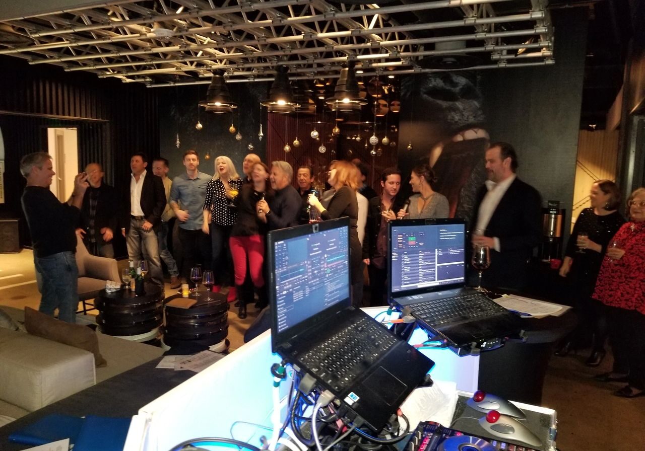 A group of people in semi-formal attire gather and socialize at an indoor event space, with DJ equipment and laptops set up in the foreground.