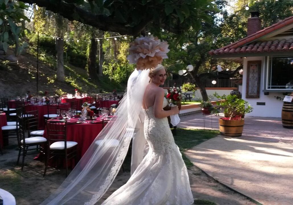 A bride in a white gown and veil holds a bouquet and smiles back while walking outdoors at a decorated wedding venue with round tables and red tablecloths.