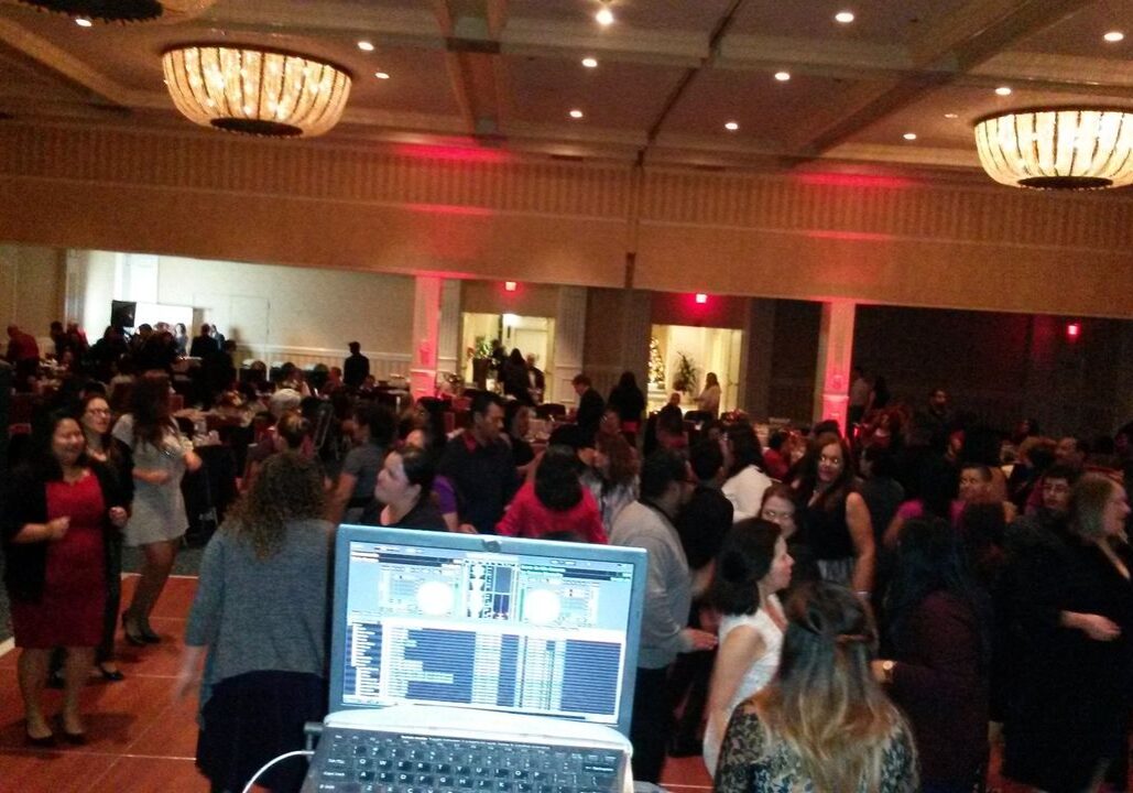 Large group of people dancing in a ballroom, seen from behind a DJ booth with a laptop displaying mixing software.