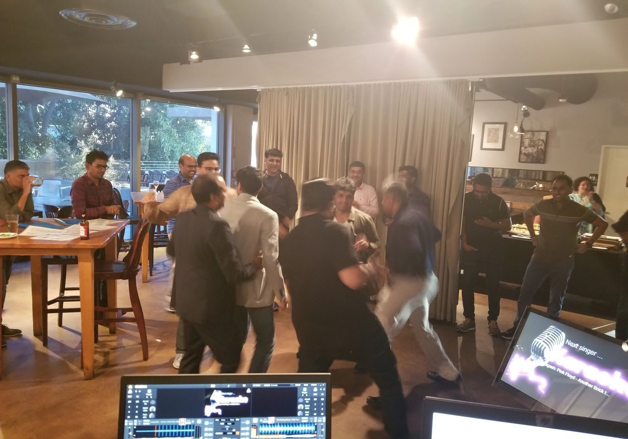 A group of people are gathered in a restaurant or cafe, some appearing to argue or confront each other while others watch. Tables, chairs, and computer monitors are visible in the foreground.