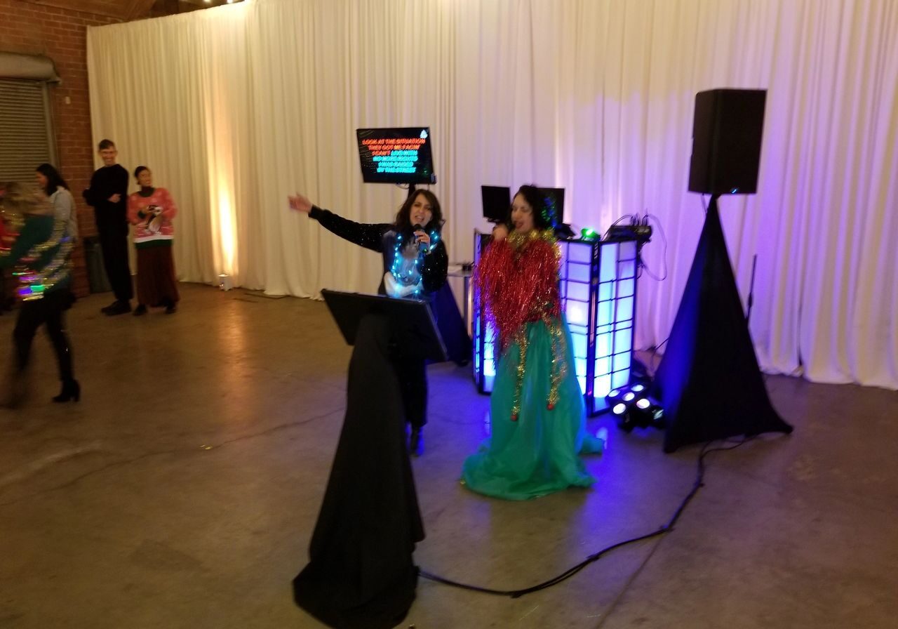 Two people sing karaoke on microphones in a decorated indoor space, with a karaoke screen, speakers, and colored lights behind them. Other people stand and watch.