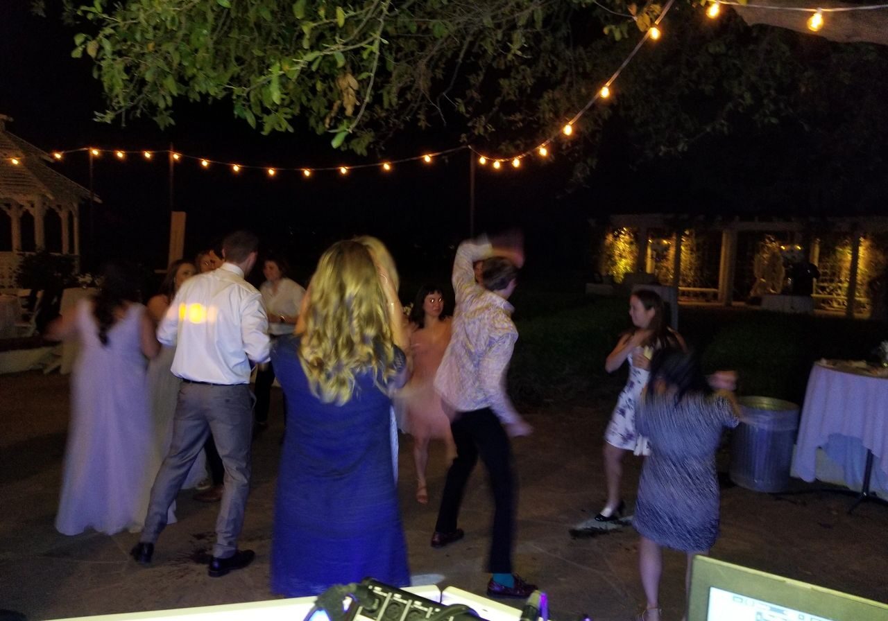 A group of people dance outdoors at night under string lights, with a DJ booth visible in the foreground and trees overhead.