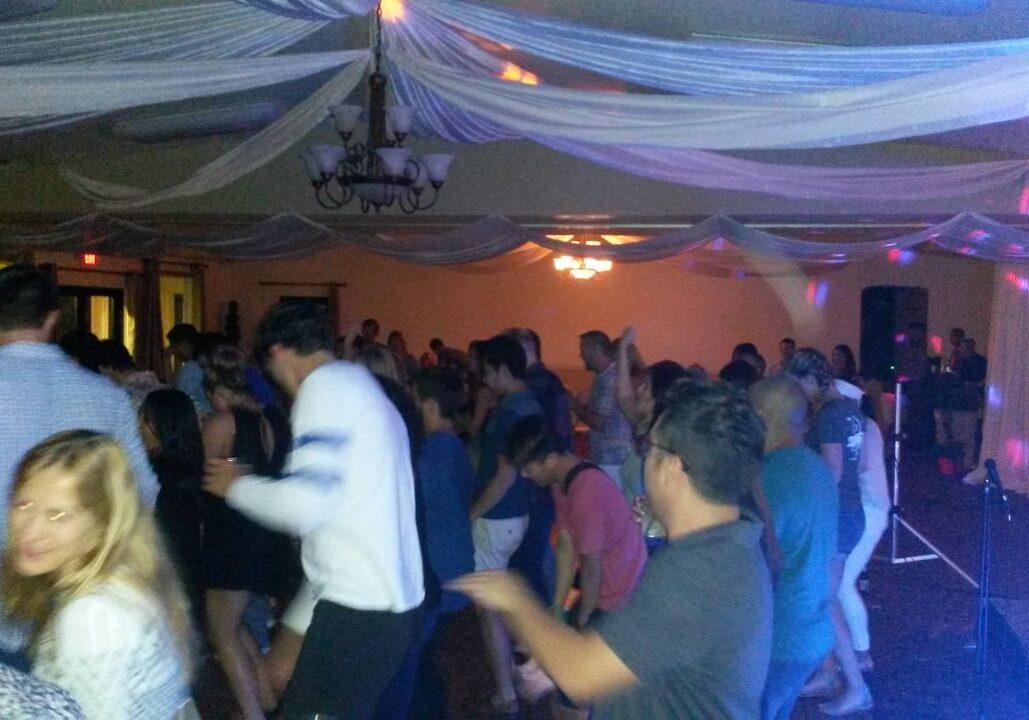 A large group of people are dancing in a decorated indoor venue with draped white fabric on the ceiling and colorful lighting effects.