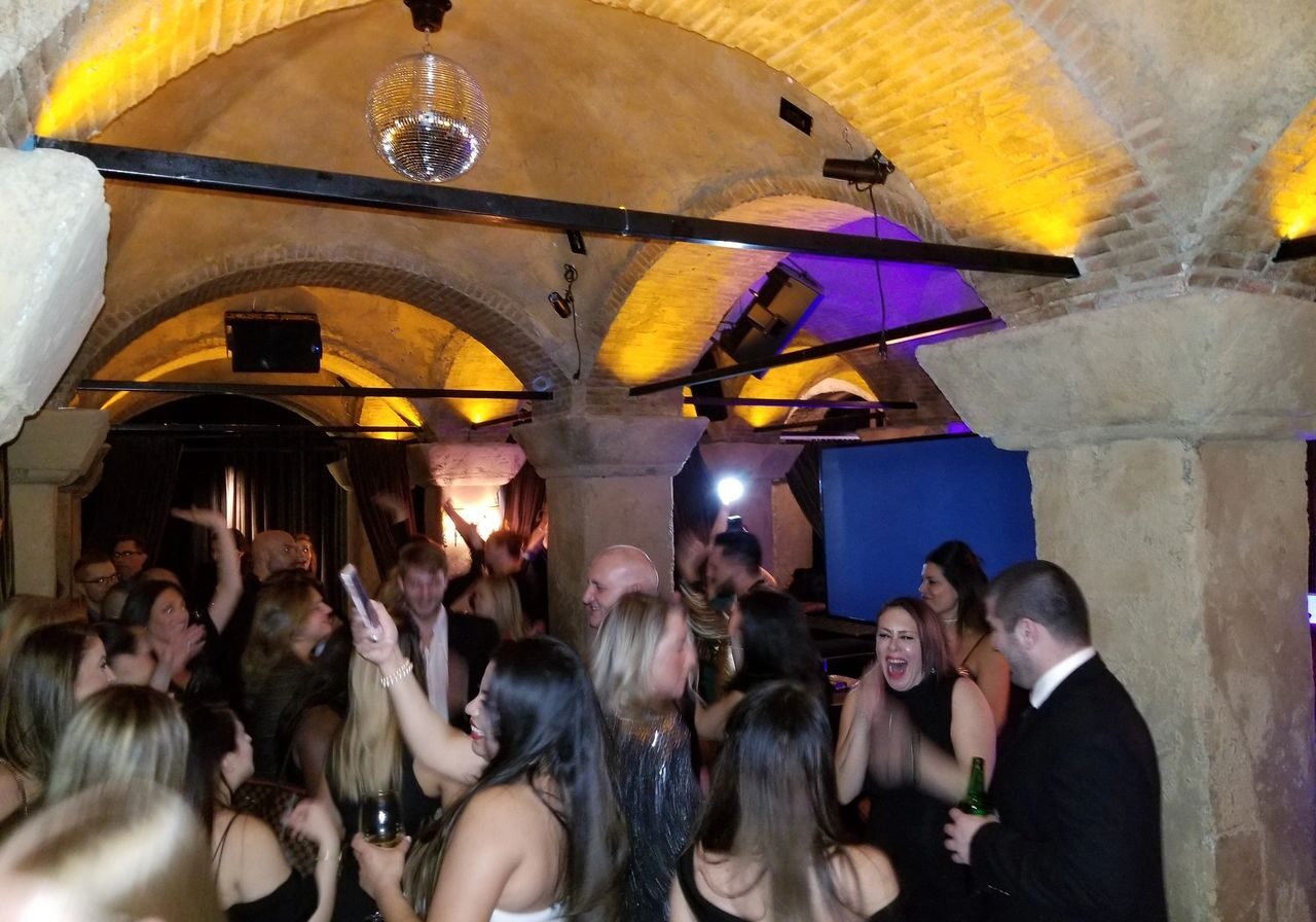 A group of people in formal attire dance and socialize under arched stone ceilings at a lively indoor party with yellow accent lighting.