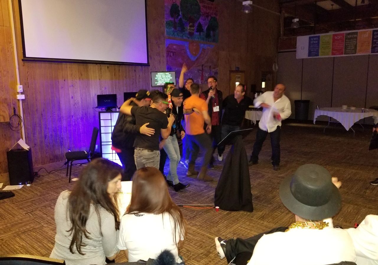 A group of people gather on a stage under a projector screen, some hugging and celebrating, while others sit and watch in a warmly lit room with wood-paneled walls.