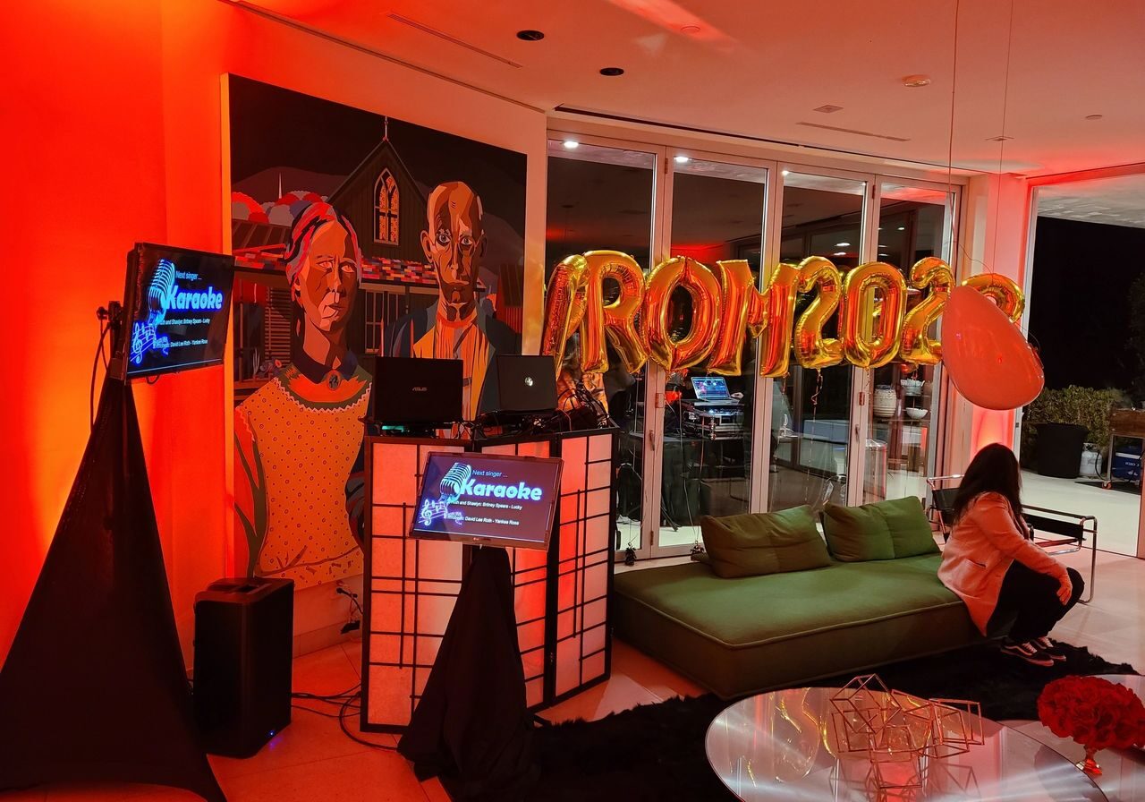 A living room decorated with "PROM 2023" gold balloons, a karaoke setup, colorful lighting, and a mural; a person sits on a green couch.