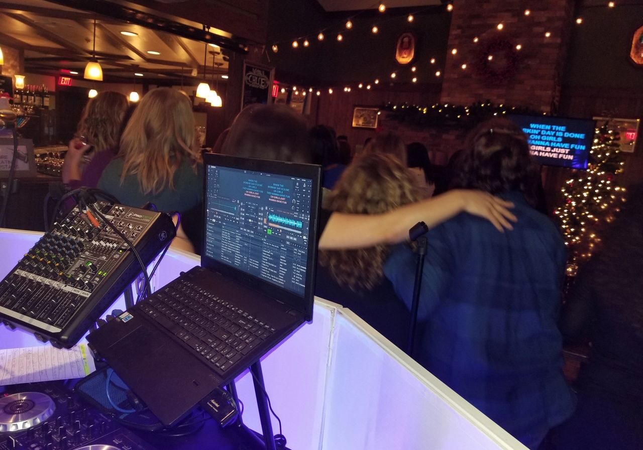 A group of people stand with arms around each other at a karaoke night in a dimly lit bar; a DJ setup and laptop are visible in the foreground.