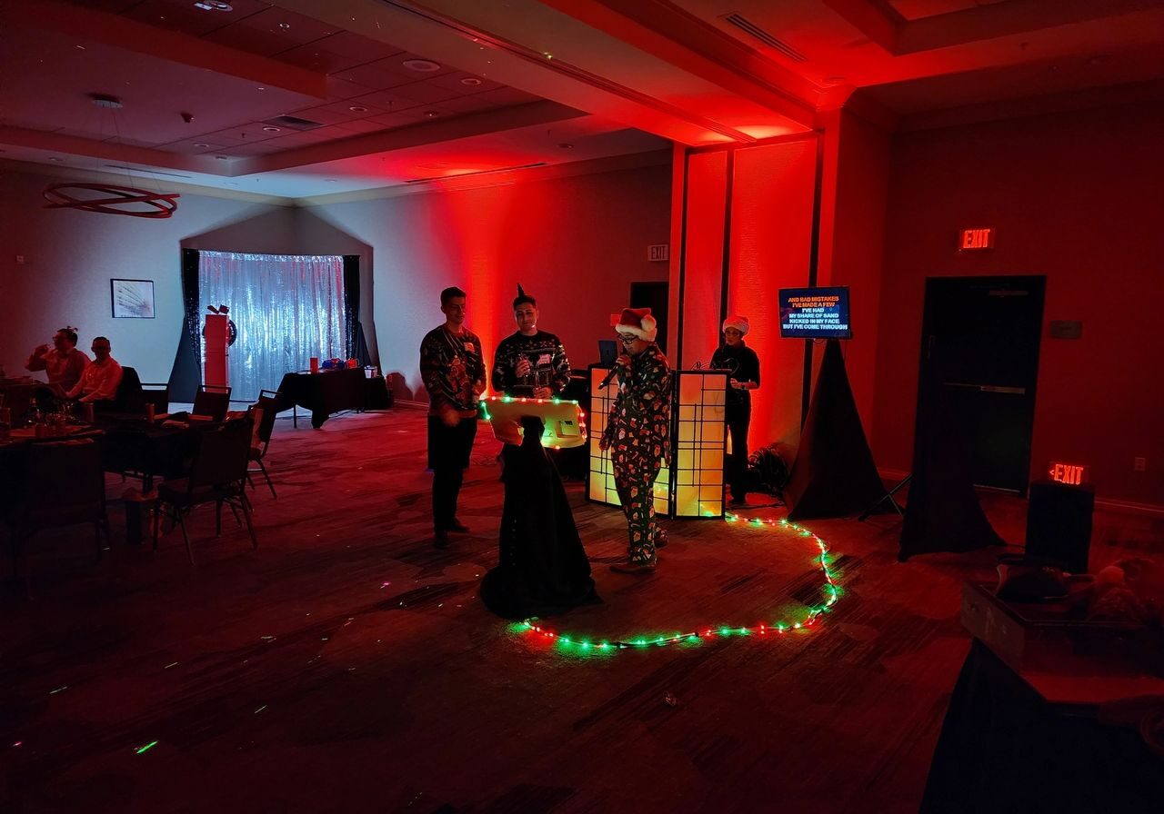 Four people in costumes stand near a lit DJ booth in a dimly lit room with red lighting and string lights on the floor; guests are seated at tables in the background.