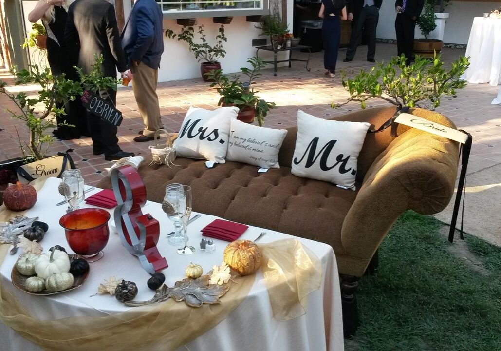 A decorated outdoor wedding table set for two with "Mrs." and "Mr." pillows on a brown loveseat, surrounded by small pumpkins and autumn-themed decor. People are standing in the background.