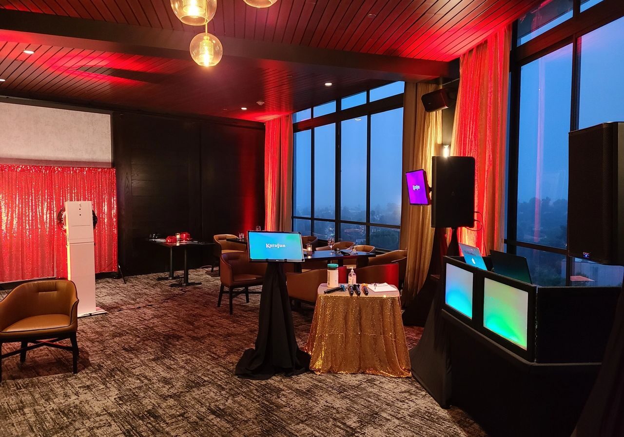 A modern lounge with floor-to-ceiling windows, red ambient lighting, small tables, chairs, a DJ booth, and digital screens set up for an event.