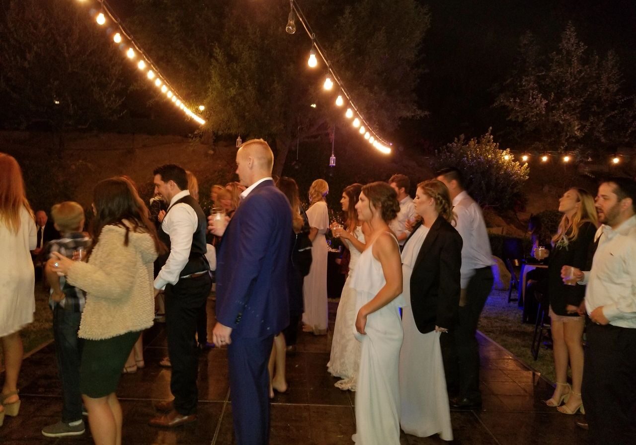 A group of people, some dressed formally, stand and dance under string lights at an outdoor nighttime event.