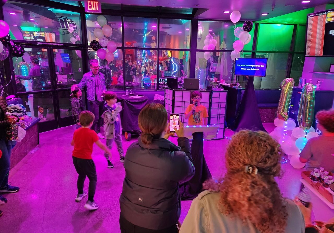 Children dance at a birthday party in a room with purple lighting, balloons, and a DJ booth while adults watch and take photos.